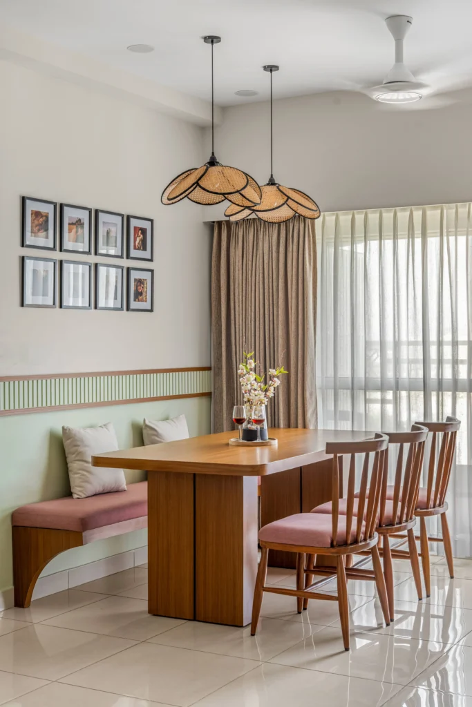 A dining room with a bold rectangular wooden table with rounded corners paired with wooden chairs featuring pinkish-red seats. On the other side, a bench is designed with the same pinkish-red seats and soft green cushions. The pair of customized pendant lights and the photo walls narrate the unforgettable memories