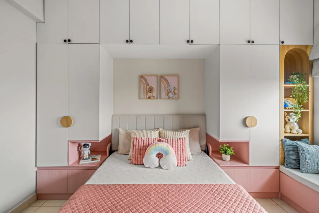 Kids' bedroom interior with a white and pink wardrobe, a personalized cozy bed with pastel cushions, a rainbow pillow, a windbay as a study nook, and decorative shelves with toys and indoor plants. A smart space-saving design is implemented with overhead storage, making it the perfect room for kids that is both stylish and functional