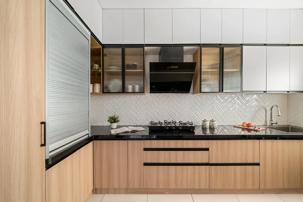 Modular kitchen interior design designed in L - shape with wooden and white cabinets. A gas stove placed on black countertop, glass-front upper cabinets, and a chimney adds elegance to the space. Herringbone white tile backsplash, under-mount sink, and minimalist handles provide a stylish and functional cooking experience