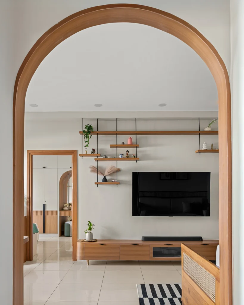 The modern living room interior of an apartment in Bangalore has a wooden arch entrance and a wall-mounted TV positioned above a chic wooden multi-purpose cabinet. The open shelves made of wood hanging from the ceiling give a unique finish to the space. Which is decorated with plants and artifacts, minimal decor, and flooring with glossy tiles